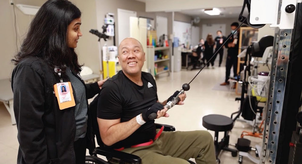 Jeff Whitaker smiles up at occupational therapist Tejaswini Thakur as he works at a weight machine 