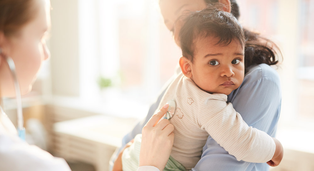 A female doctor seen from behind extends a stethoscope to a sad toddler being held by their mother.