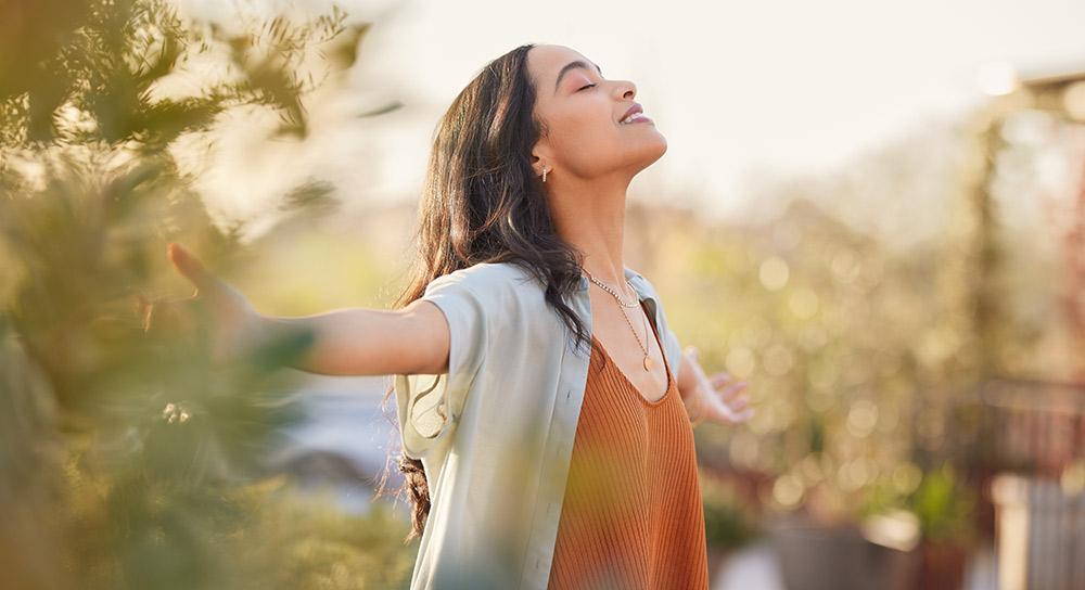 A young woman with brown hair, wearing an orange sweater and gray cardigan, holds her arms out wide and looks up with eyes closed at the sky