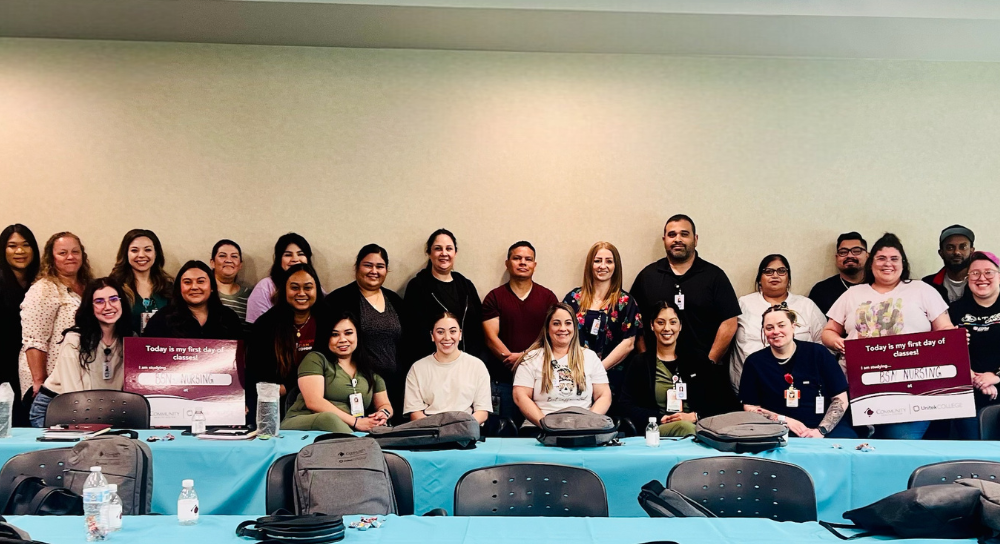 A group of about 20 people, all genders, pose together against the wall of a classroom