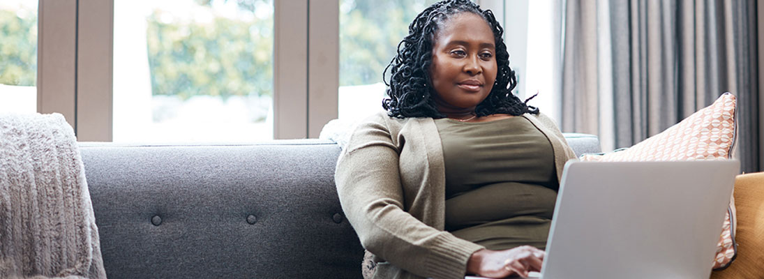 An African American woman sits on her couch looking interestedly at her laptop computer.