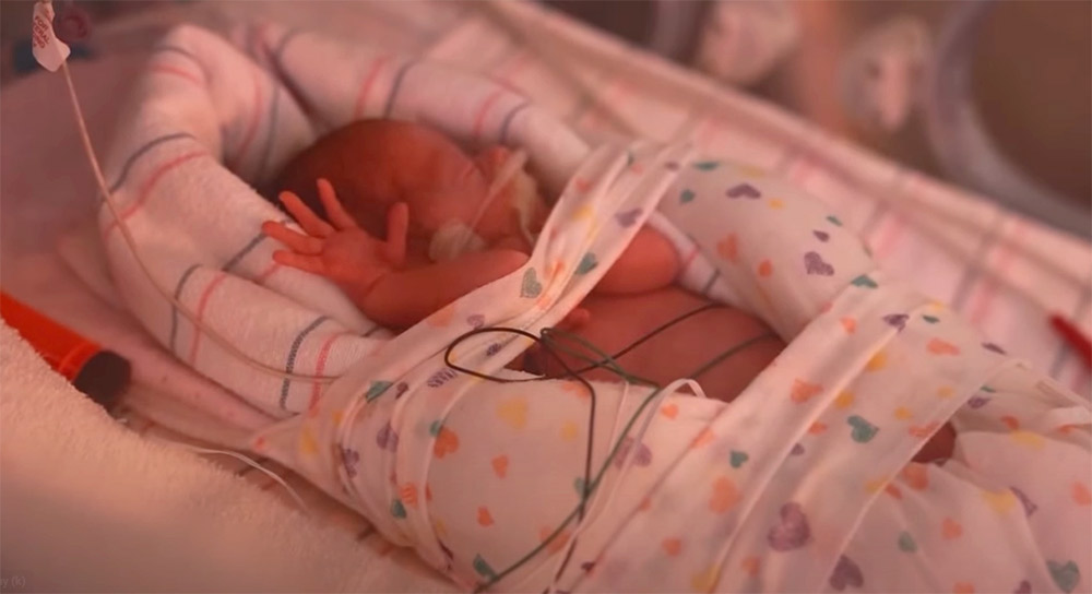 A tiny baby reaches out a hand from beneath blankets in an an incubator.