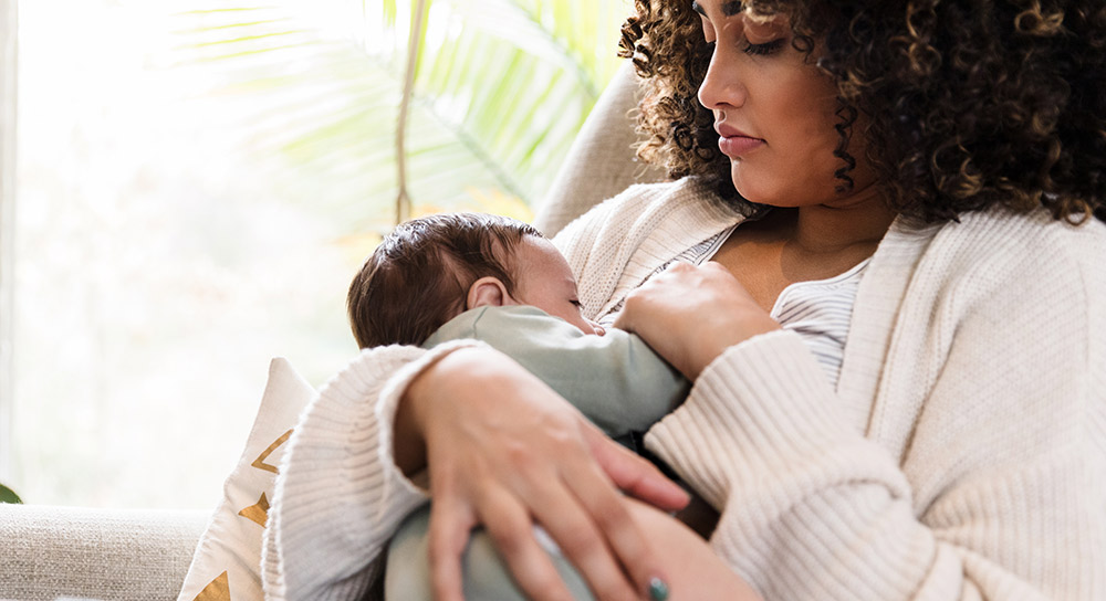 A young, Black mother sits in a comfortable chair and holds her small baby close to her chest. 