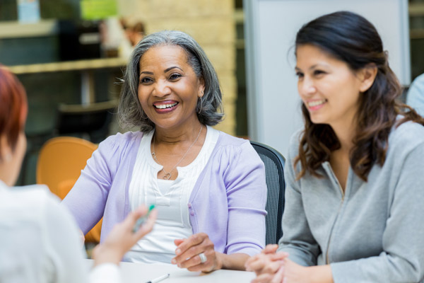 Two women sit in a support circle smiling. The woman on the left is Black with grey hair; the woman on the left is white with brown hair. 