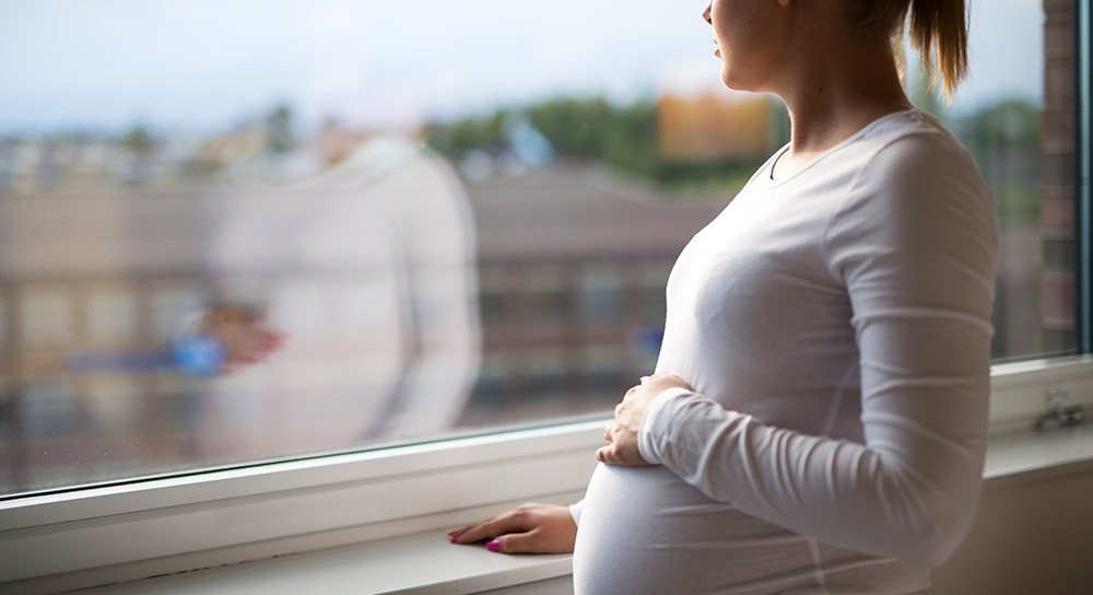 A young white mother touches her stomach and stares through a window into the distance.