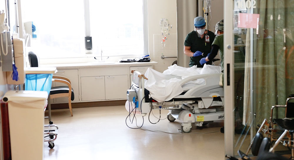 Looking into a hospital room, a provider in blue scrubs and mask is tending to an unseen patient.