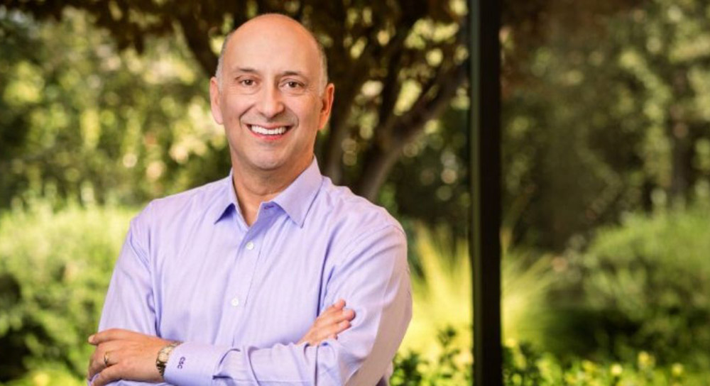 Craig Castro stands with arms crossed in a light purple long-sleeve shirt in front of a green nature background