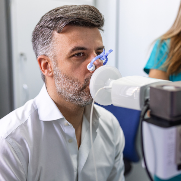 A white woman with blond hair sits upright in a hospital bed and breathes into a lung diagnostic tool