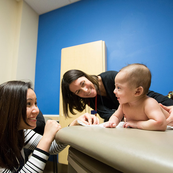 A young mother smiles at her baby, who is being held by a healthcare worker in an exam room